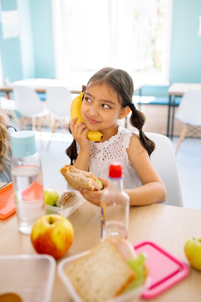 A young girl playfully holds a banana while enjoying a sandwich at a classroom table. Kindergarten